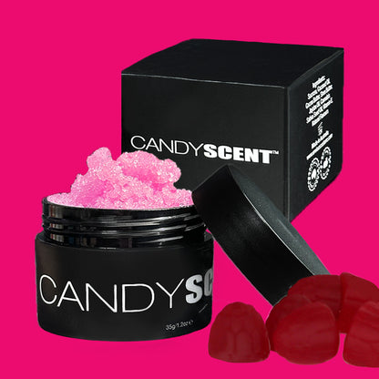 A black jar labeled "(DISCONTINUED CANDYSCENT™) Lip Sugar Scrubs," filled with pink granules, is open with its lid leaning against it. Behind the jar stands a matching black box. Red gummy candies are positioned in the lower right corner. The bright pink background enhances this tempting lip-smacking experience of our CANDYSCENT™ Lip Sugar Scrub for soft lips.