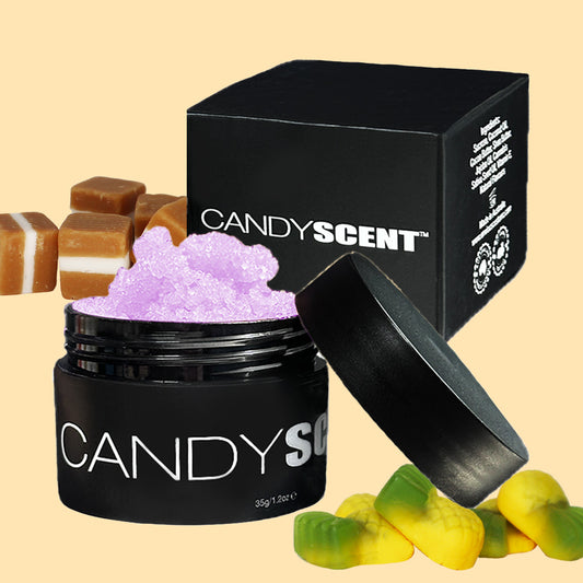 A black container labeled "CANDYSCENT™" reveals the purple Lip Sugar Scrub, infused with nourishing oils, surrounded by cubes of caramel candy and green-yellow candy pieces. In the background, there is a closed black box with the same branding. The light beige backdrop highlights this inviting scene.