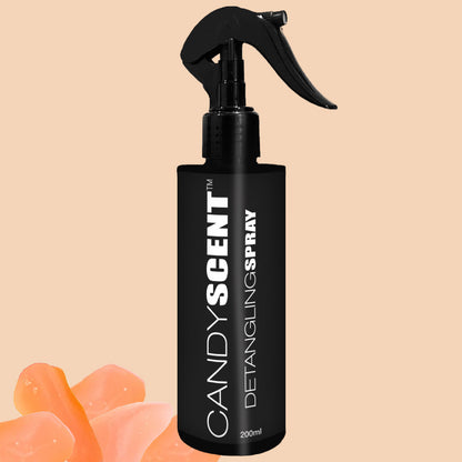 A black spray bottle labeled "CANDYSCENT™ Hair Detangling Spray" with a trigger nozzle, containing 200ml of product and infused with aloe vera and jojoba oil, rests against a soft peach background. Semi-transparent peach-colored soap pieces are positioned at the bottom left.