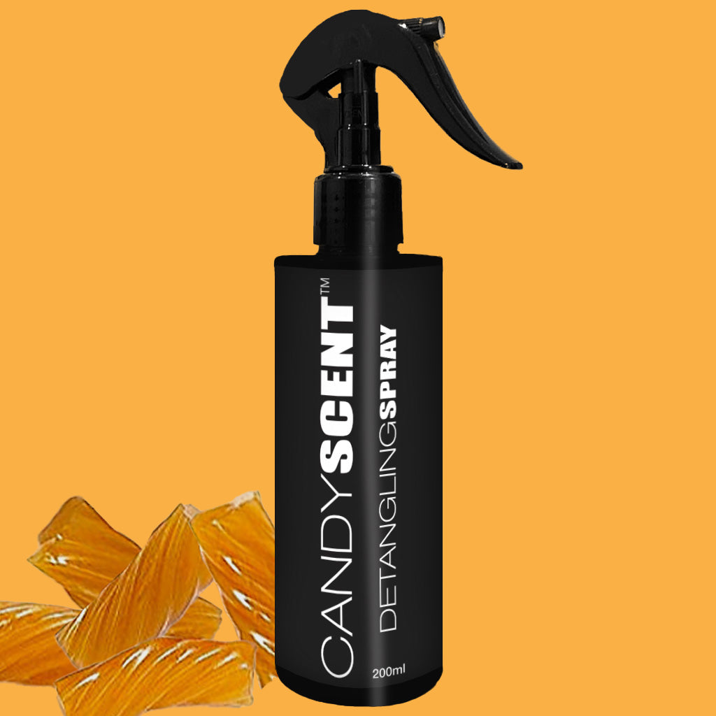 A black 200ml spray bottle of "CANDYSCENT™ Hair Detangling Spray" stands out against a vivid orange background. The bottle features a spray nozzle, and orange candy pieces are positioned in the bottom left corner. Infused with Aloe Vera and Jojoba Oil, it is ideal for all hair types.