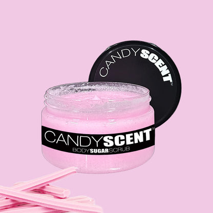 A jar of CANDYSCENT™ Body Sugar Scrub with a black lid slightly open. The scrub inside the jar is a light pink color, matching the pastel pink background. Pink, stick-like applicators are positioned next to the jar. Infused with nourishing oils, this scrub promises to exfoliate and hydrate your skin.