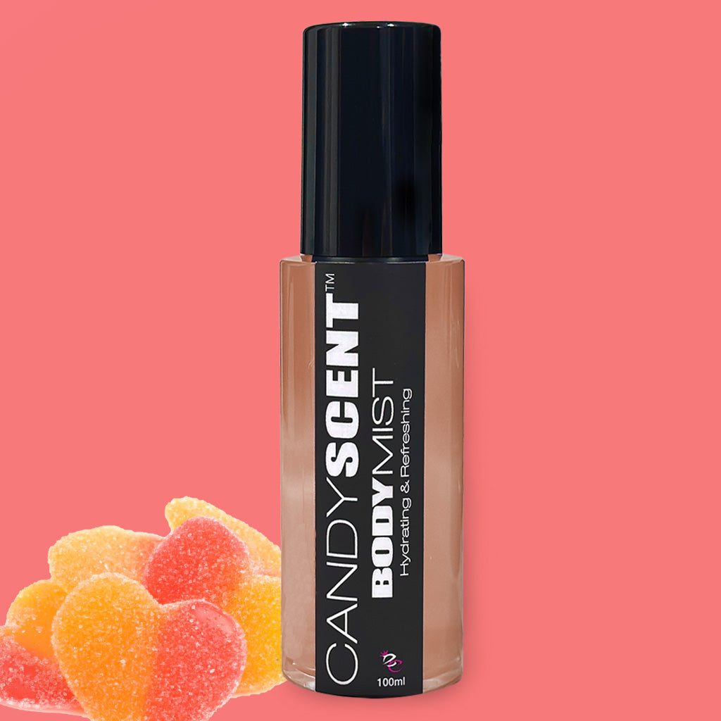 A bottle of CANDYSCENT™ Hydrating Body Mist with a black cap against a pink background. The label reads "Hydrating & Refreshing." In the foreground, three heart-shaped gummy candies in red, orange, and yellow colors are displayed.