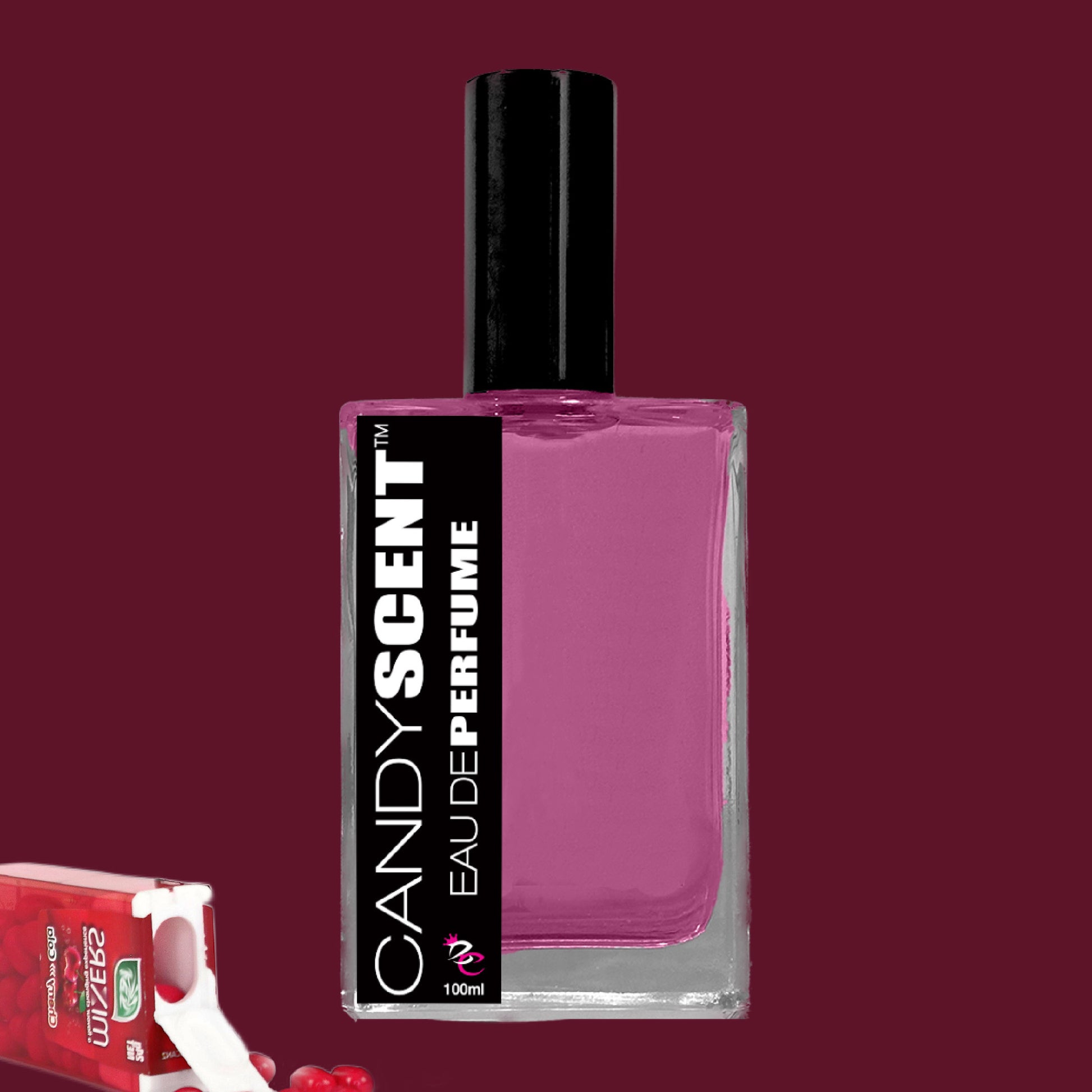 A rectangular bottle of CANDYSCENT™ Eau De Perfume / Cologne with a black cap and purple liquid inside. The black and white label details the brand and product name. A partially opened red candy wrapper with sugary treats is visible in the bottom left corner, evoking sweet playful fragrances.