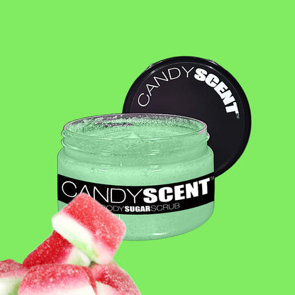 A jar of CANDYSCENT™ Body Sugar Scrub with a black lid lies open, revealing green scrub infused with nourishing oils inside. The label reads "CANDYSCENT™ Body Sugar Scrub." Pink and white candy pieces are placed in the bottom left corner. The background is a vibrant green.