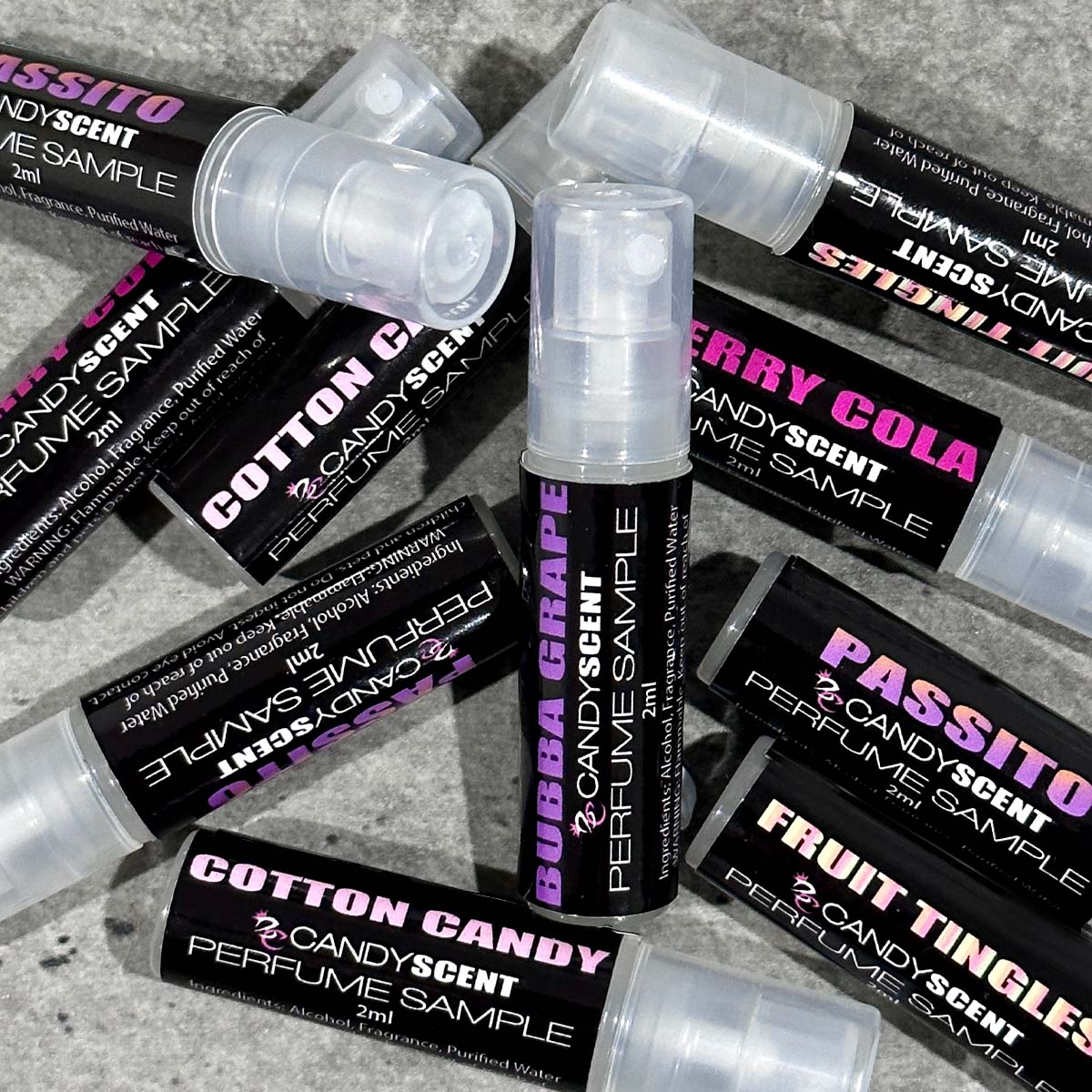 A scattered pile of CANDYSCENT™ Eau De Perfume / Cologne sample vials. Each vial has a black label with bright text highlighting various scents, such as "Cotton Candy," "Bubba Grape," and "Berry Cola." These sweet, playful fragrances are reminiscent of sugary treats. The vials are on a gray surface.