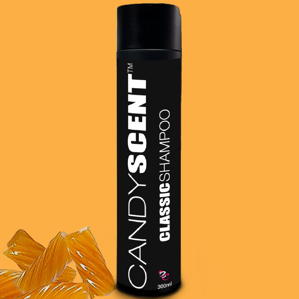 A black bottle of CANDYSCENT™ Classic Shampoo stands upright against an orange background. The luxurious shampoo, labeled "Classic Shampoo 300ml" under the CANDYSCENT™ brand, is accompanied by scattered orange hard candies in front of the bottle.