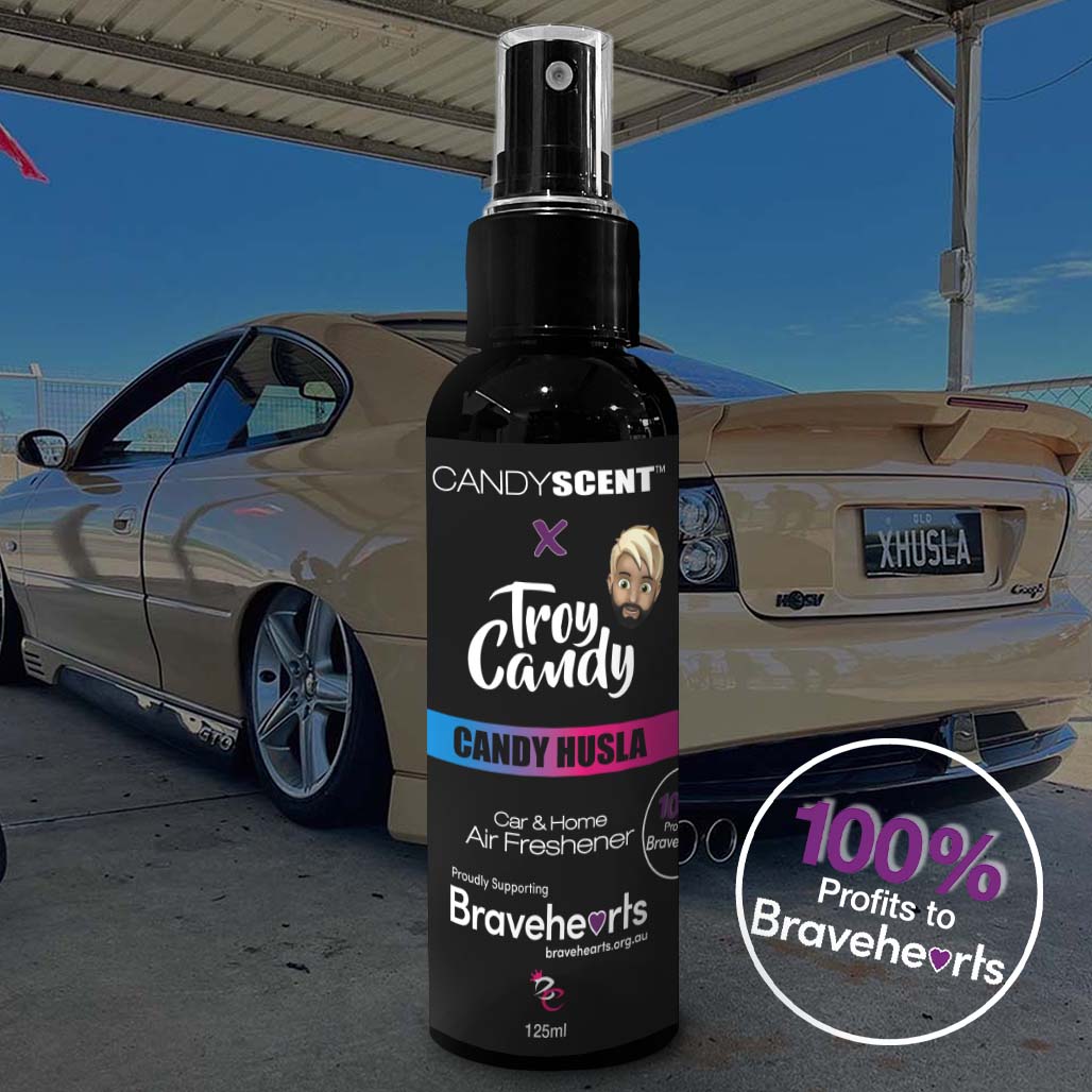 A CANDYSCENT™ x Troy Candy Car Spray bottle, branded under the name CANDYSCENT™, featuring the delightful Candy Husla scent, is prominently displayed in the foreground. Behind it, a sleek beige sports car rests under a roof. The bottle proudly states, "100% of Profits to Bravehearts Foundation.