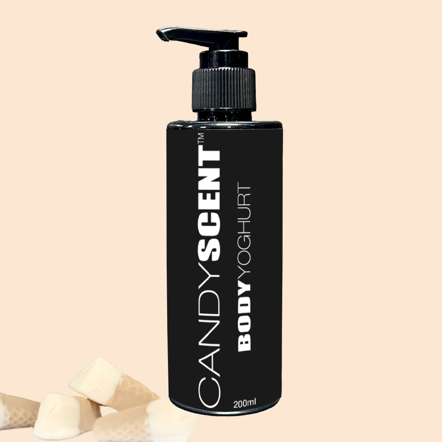 A black bottle of Body Yoghurt by CANDYSCENT™ with a pump dispenser sits elegantly against a beige background. Three small pieces of white chocolate rest at the bottom left corner. The label, prominently showing "200ml," promises a nourishing experience and deep hydration for your skin.