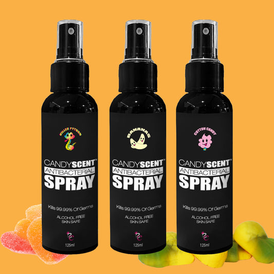 Three black bottles of CANDYSCENT™ Antibacterial Spray are shown against a solid orange background. The sprays, labeled with scents like Killer Python, Banana, and Cotton Candy, are alcohol-free and kill 99.99% of germs. They are flanked by colorful gummy candies and a few flower petals.