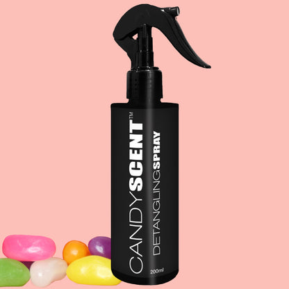 A black bottle of CANDYSCENT™ Hair Detangling Spray with a trigger nozzle is showcased against a pastel pink background. Infused with aloe vera and featuring a delightful candy scent, this detangler is surrounded by six colorful jellybeans in shades of pink, white, yellow, green, and purple.