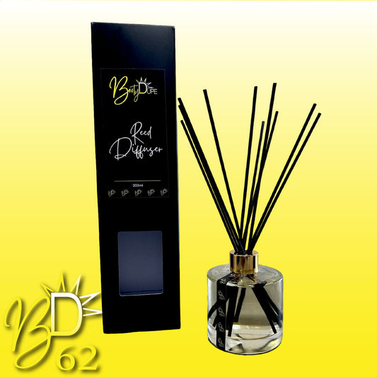 A black reed diffuser box labeled "Reed Diffuser" from BOOTY DUPE stands next to a glass diffuser jar with black reeds fanning out. The background transitions from yellow to white, featuring a yellow "BD 62" logo with a sunburst design in the bottom left corner. This Australian-made BOOTY DUPE reed diffuser promises long-lasting fragrance.