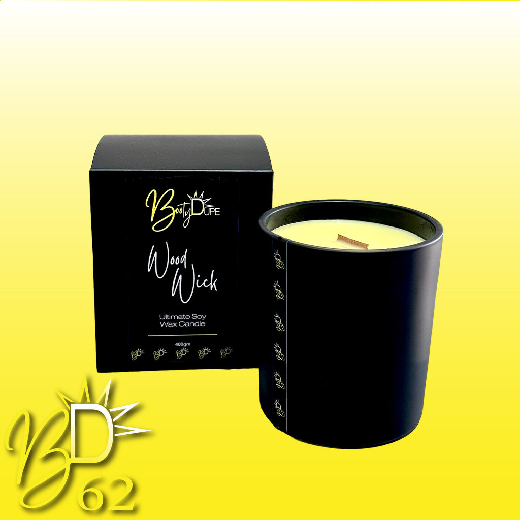 A luxurious, hand-poured Wood Wick Soy Candle in a black glass container with a wooden wick is showcased next to its sleek black box packaging. Both the candle and the box prominently feature the BOOTY DUPE brand name and a logo with the initials "BD." The background features a gradient from yellow to white.