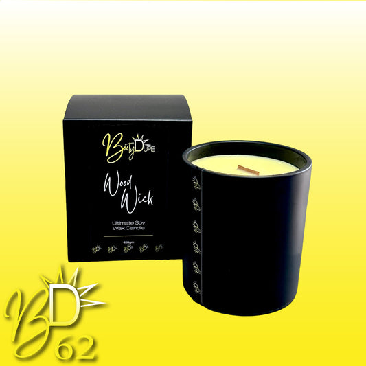 A luxurious, hand-poured Wood Wick Soy Candle in a black glass container with a wooden wick is showcased next to its sleek black box packaging. Both the candle and the box prominently feature the BOOTY DUPE brand name and a logo with the initials "BD." The background features a gradient from yellow to white.